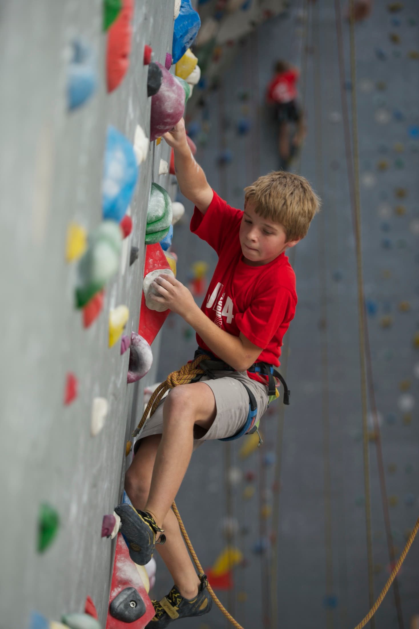 Young Leto on an indoor rope wall in a red t-shirt, tied in and wearing a harness