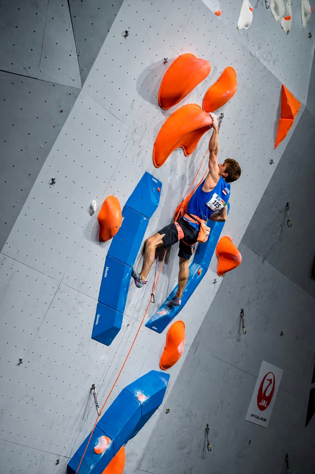 Reaching for an orange hold on a grey competition lead wall, wearing a Netherlands singlet