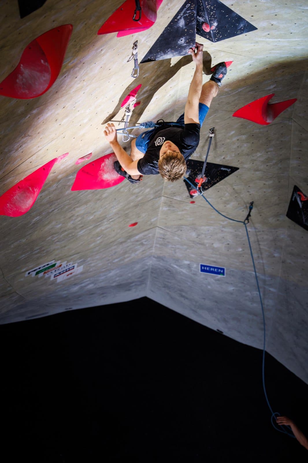 Clipping the rope on a steep plywood overhang covered in red holds, seen from above