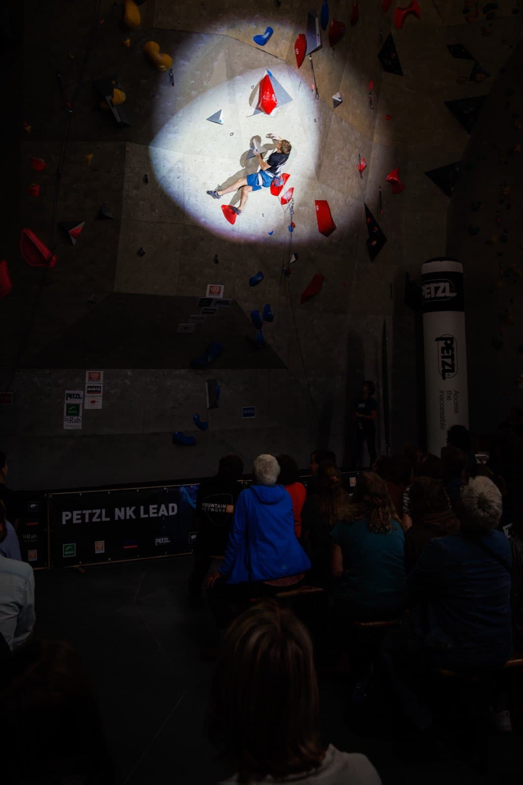 Spotlit high on a lead wall in a darkened hall, an audience watching from below