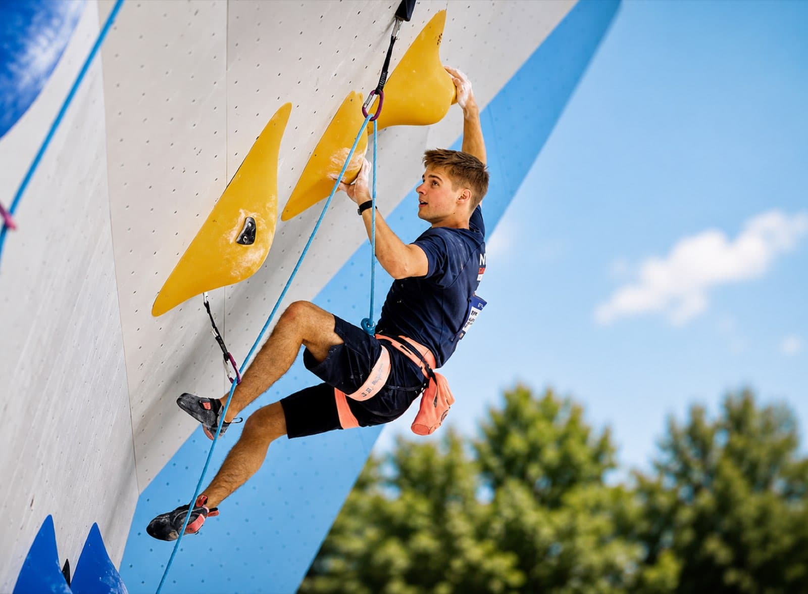 Hanging from a yellow hold on an outdoor competition wall against a blue sky and trees