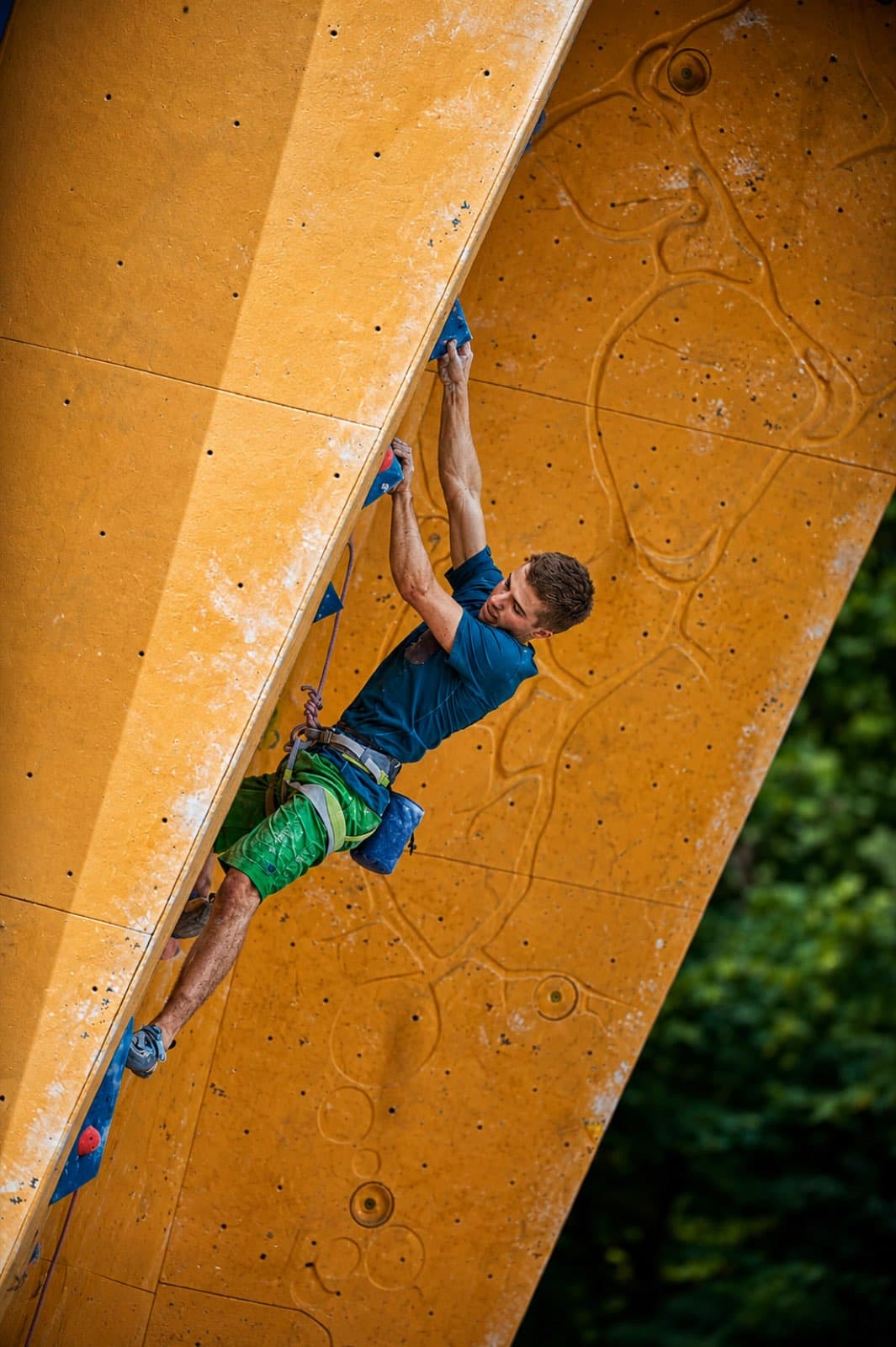 Lead climbing an overhanging orange and yellow competition wall outdoors, forest behind