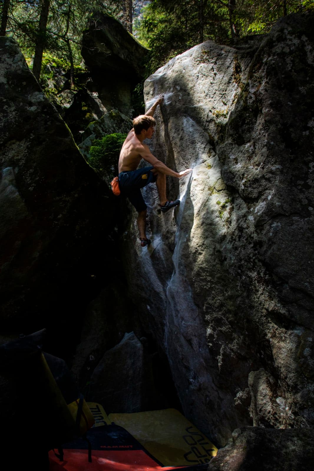 Topping out a forest boulder on a steep grey arête, crash pads below