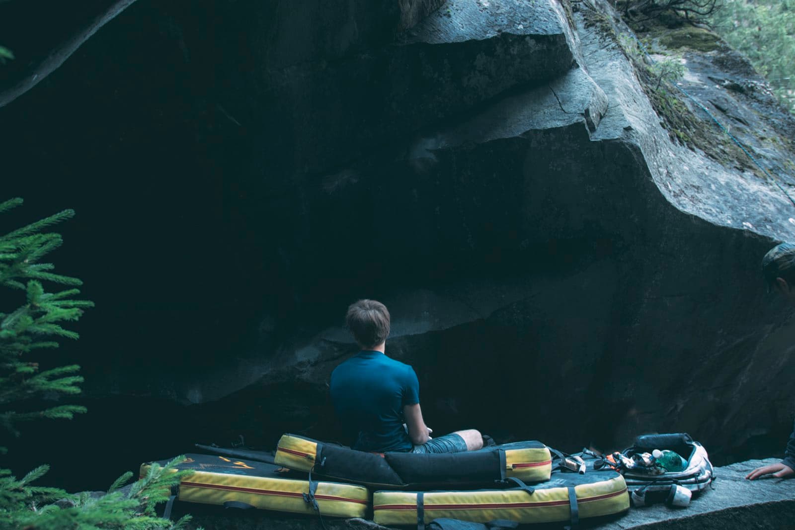 Sitting on crash pads below a large boulder in the forest, looking up at the line