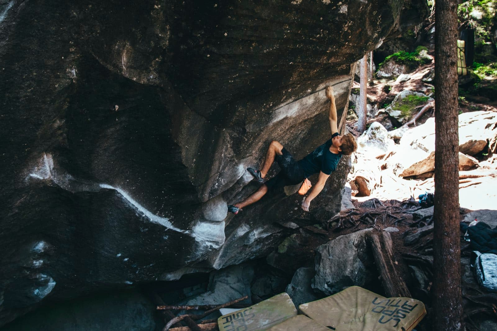 Bouldering out a granite roof in the forest, sunlit rocks behind