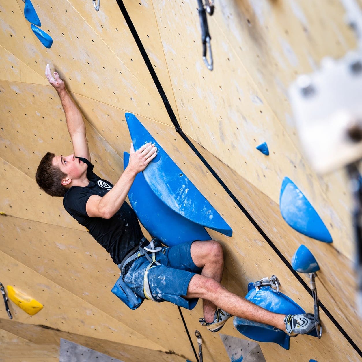 Reaching high off a blue volume on an overhanging plywood competition wall, the rope trailing below