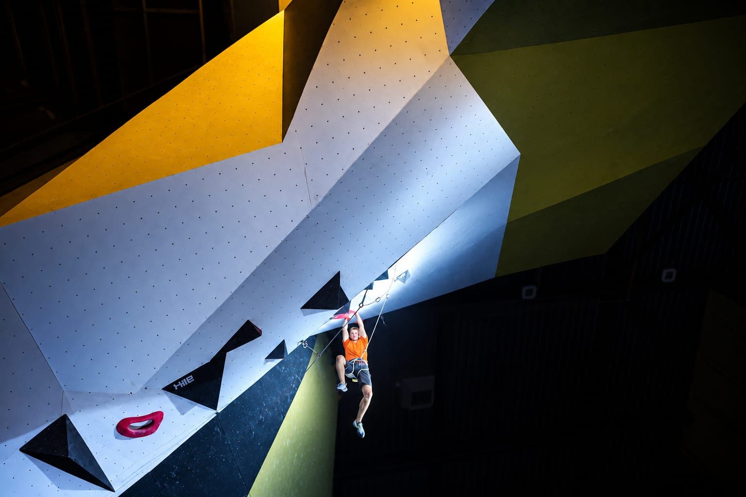 Out on the lip of a steep competition roof under a spotlight, clipping high above a dark hall