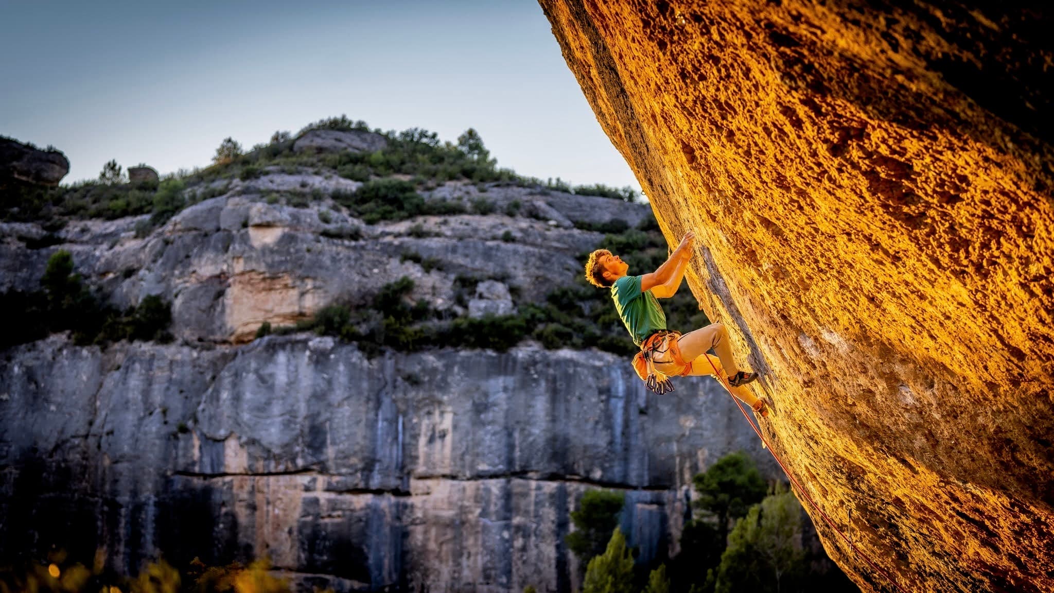 Climbing an overhanging orange limestone wall at Margalef, the canyon wall and trees behind