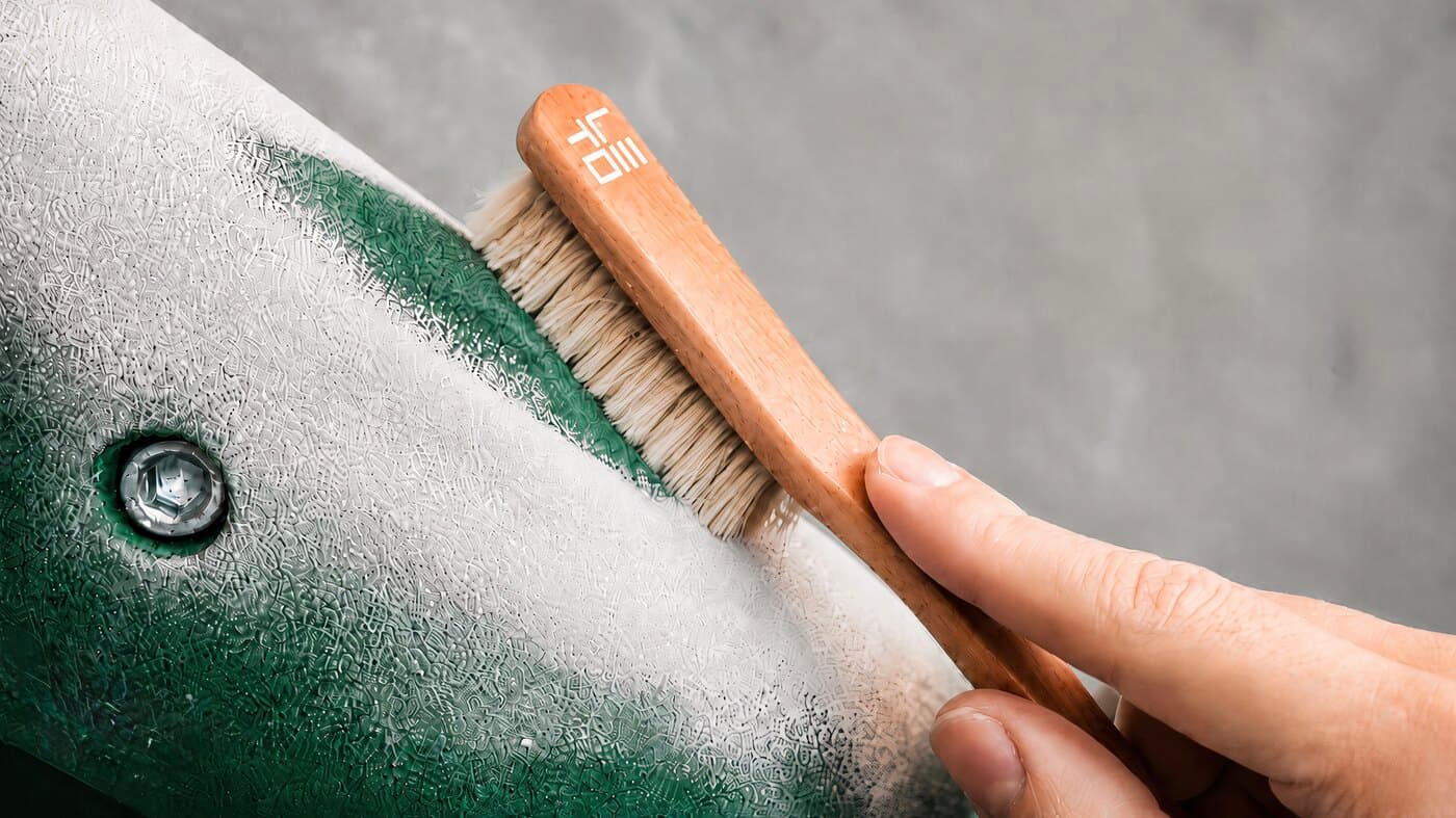 Een hand die de houten borstel met logo tegen een groen-witte klimgreep houdt en er het magnesium afveegt