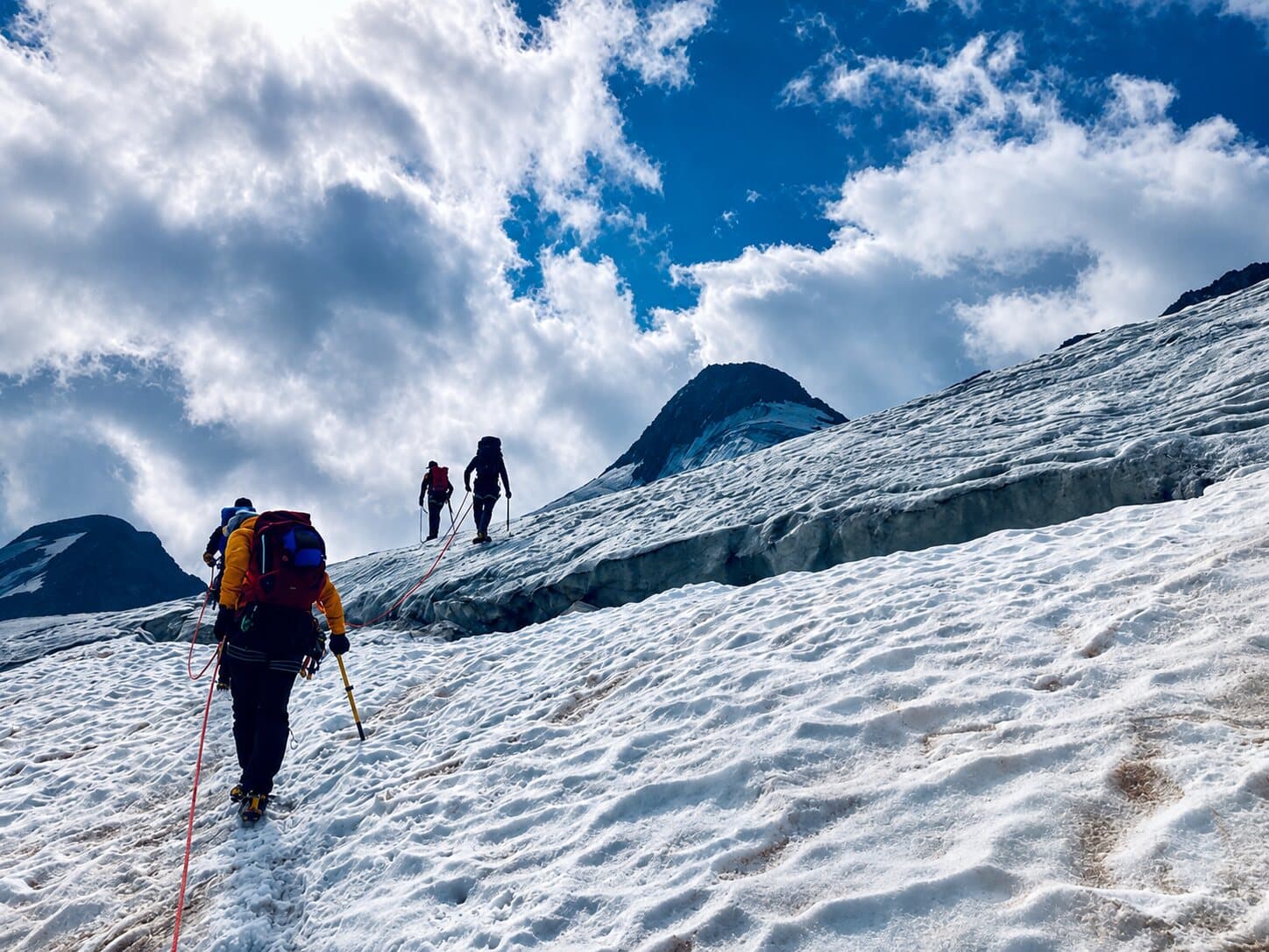 A roped team of three walking up a snow slope on a glacier, a rock peak ahead under broken cloud