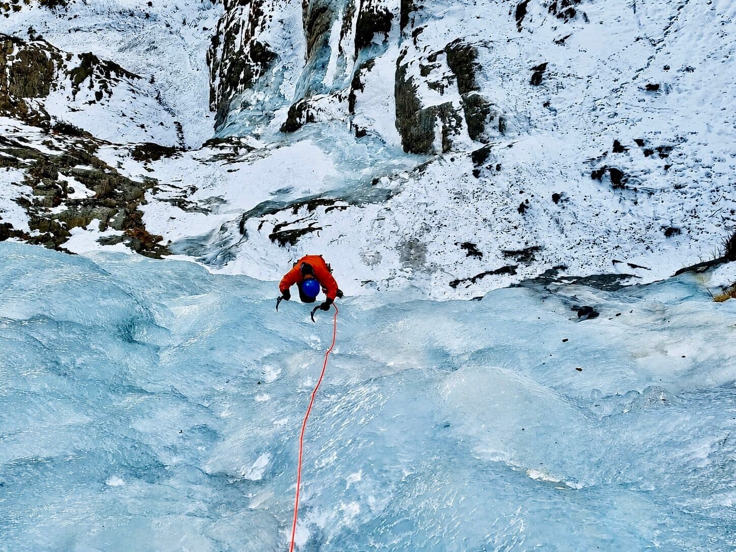 The same frozen line seen from further back: a climber small on the blue ice below snow-covered rock bands