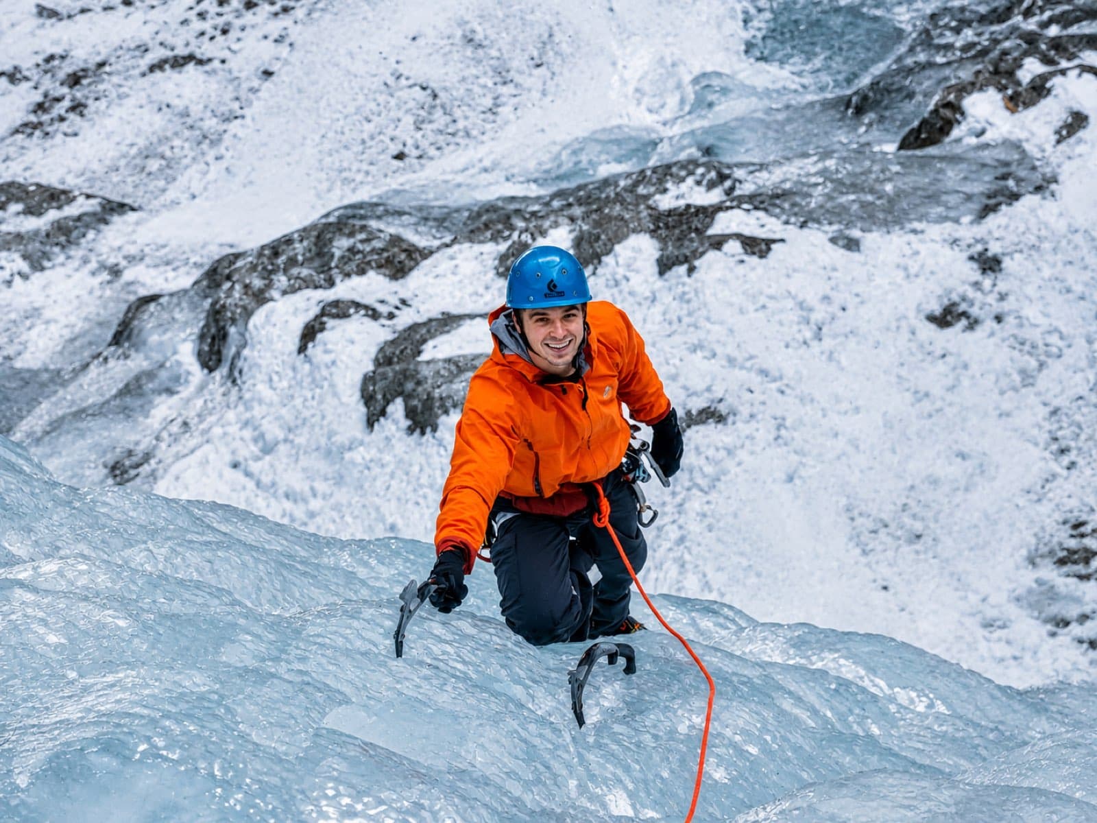 Climbing a wall of blue glacial ice with two ice tools, wearing an orange jacket and blue helmet