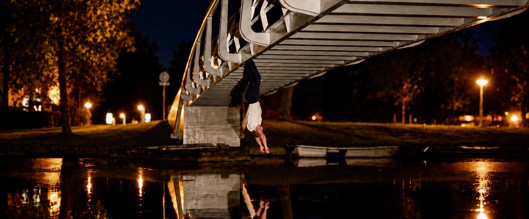 's Nachts ondersteboven aan de voeten hangend aan de ligger van een gebogen voetgangersbrug, armen omlaag, de brug en de figuur allebei weerspiegeld in stil water