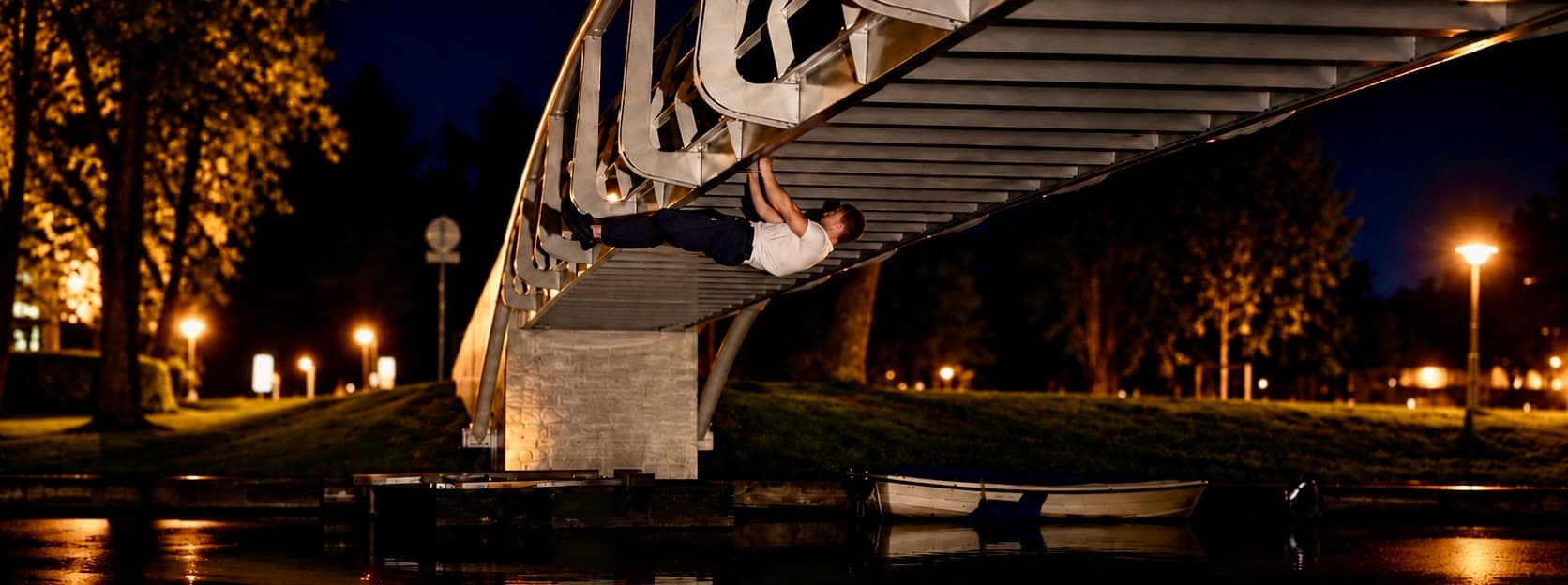 's Nachts een front lever onder het dek van een gebogen voetgangersbrug over een gracht, het lichaam horizontaal, lantaarnlicht langs beide oevers en een afgemeerde boot eronder
