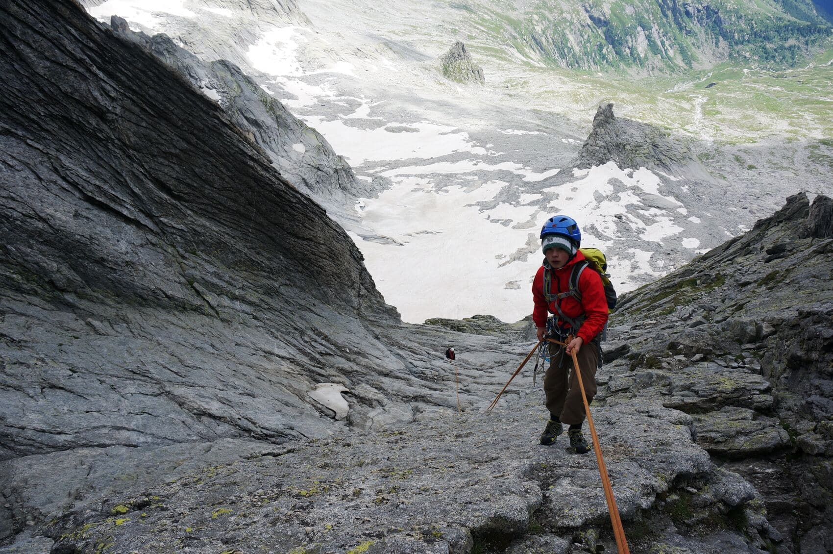A young climber in a red jacket, helmet and pack on the granite of Piz Badile, a snowfield and the valley far below