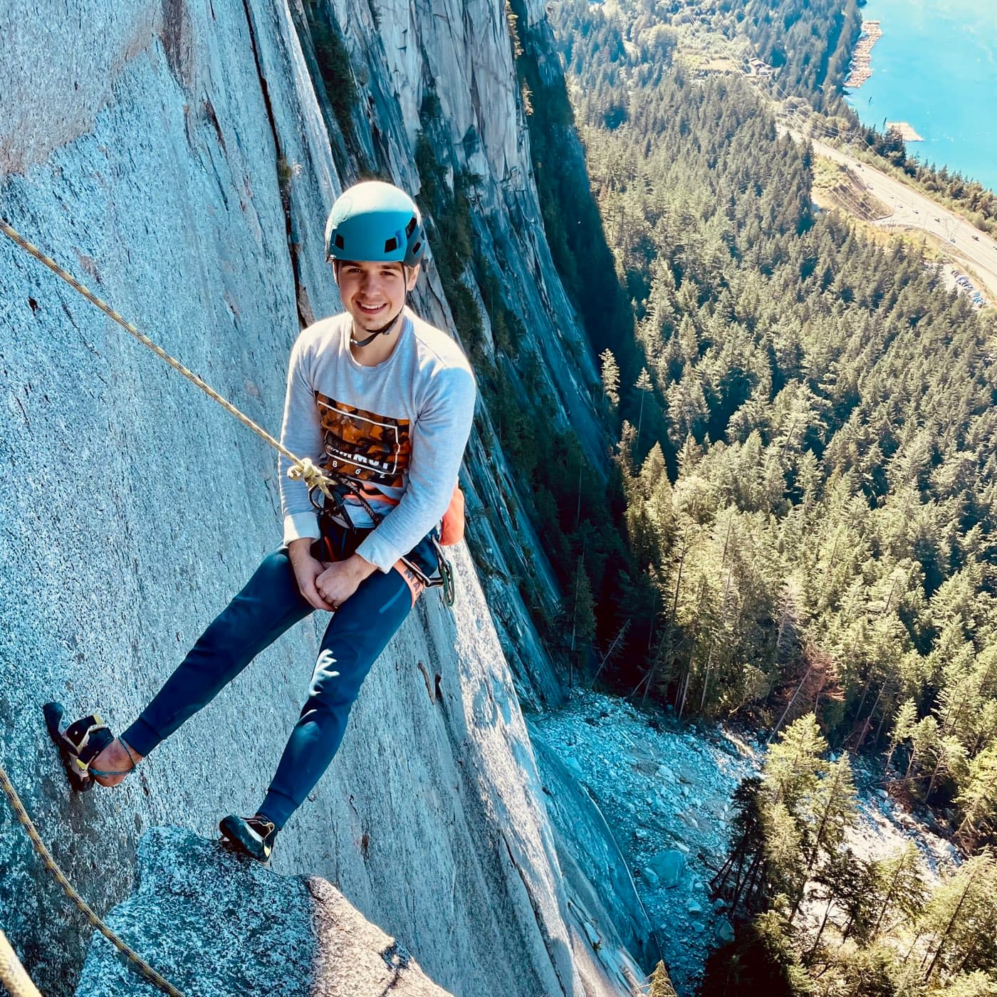 Climber at a hanging belay high on a granite wall in Squamish, forest and inlet far below