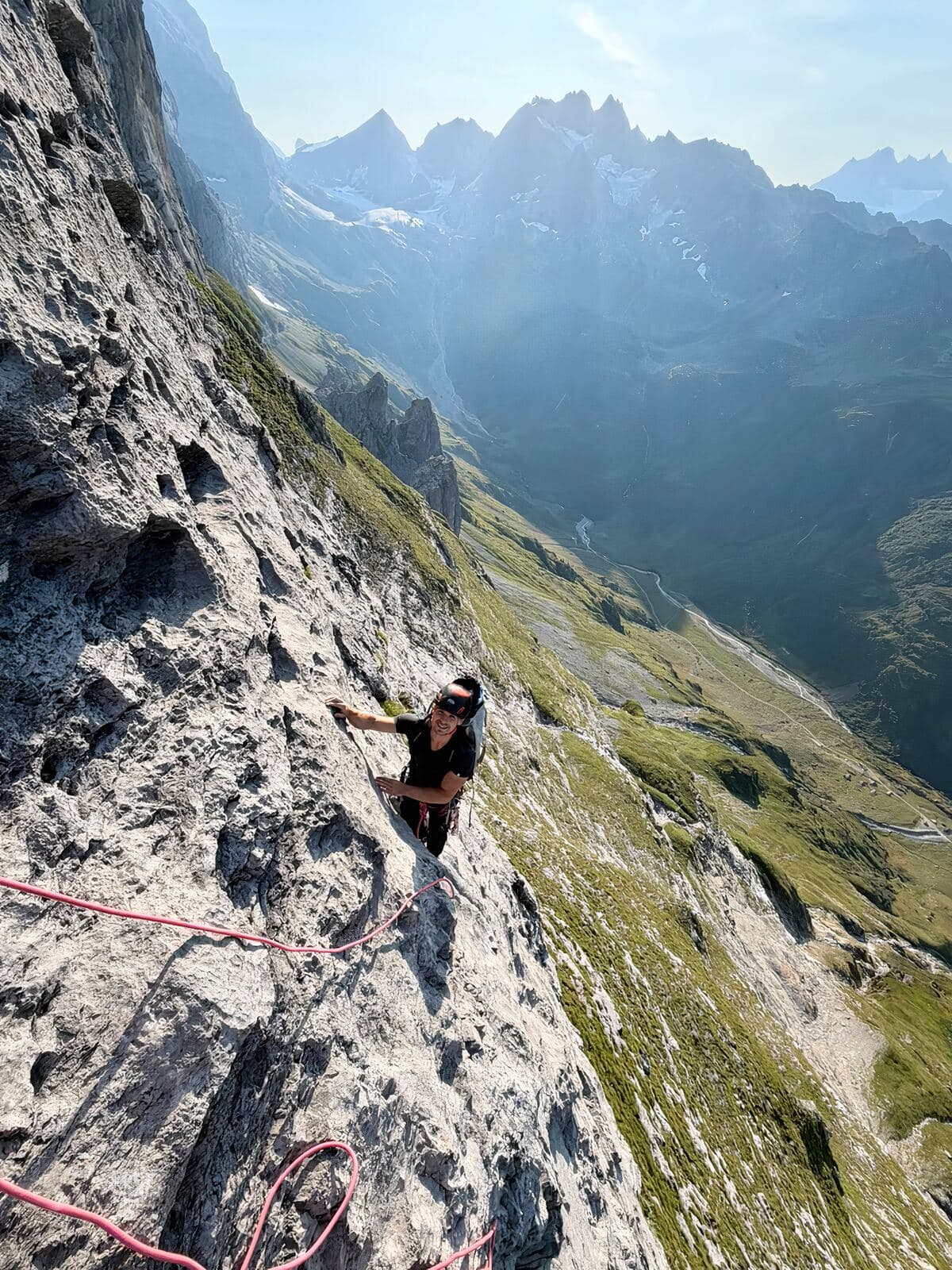 A climber on the wall looking back across the valley, snow-covered peaks behind and ropes running through the foreground